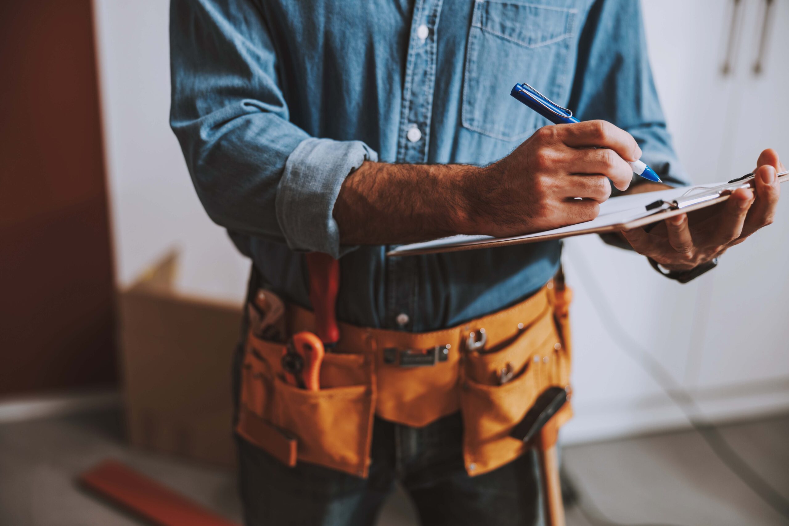 Construction worker in a denim shirt writes on a clipboard with a blue pen.